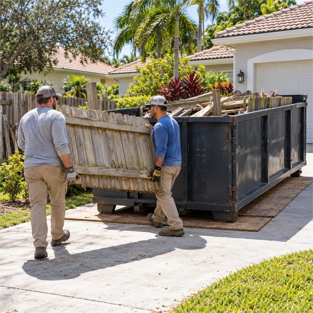 Two workers carrying an old wooden fence panel toward a roll-off dumpster filled with demolition debris during dumpster rental services near me