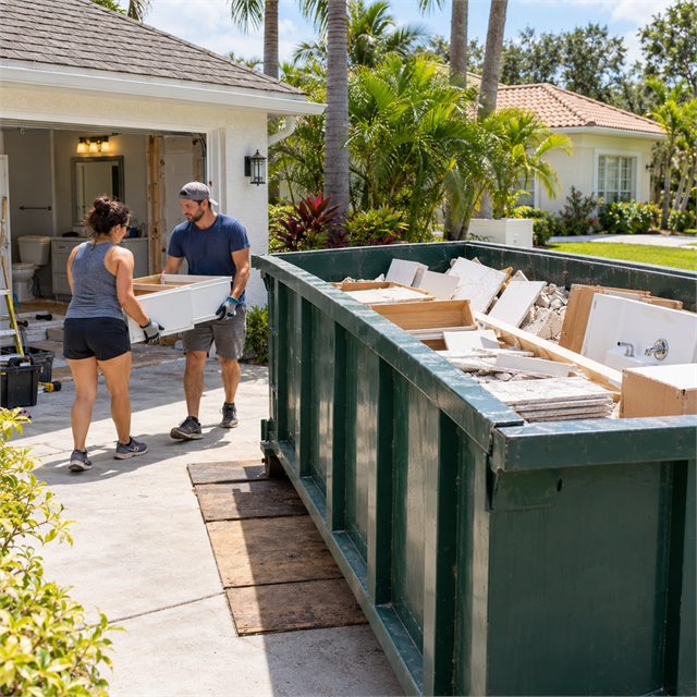 Two workers carrying a bathroom vanity toward a roll-off dumpster filled with remodeling debris during dumpster rental services near me